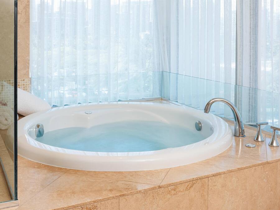 A large, oval-shaped hot tub in a modern bathroom with a glass partition and a view of tall buildings.