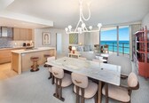 A modern kitchen with a marble island, a dining area with a large table, and a balcony with a view of the ocean.