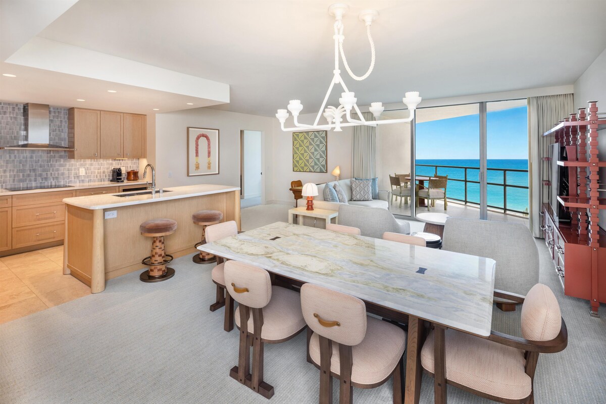 A modern kitchen with a marble island, a dining area with a large table, and a balcony with a view of the ocean.