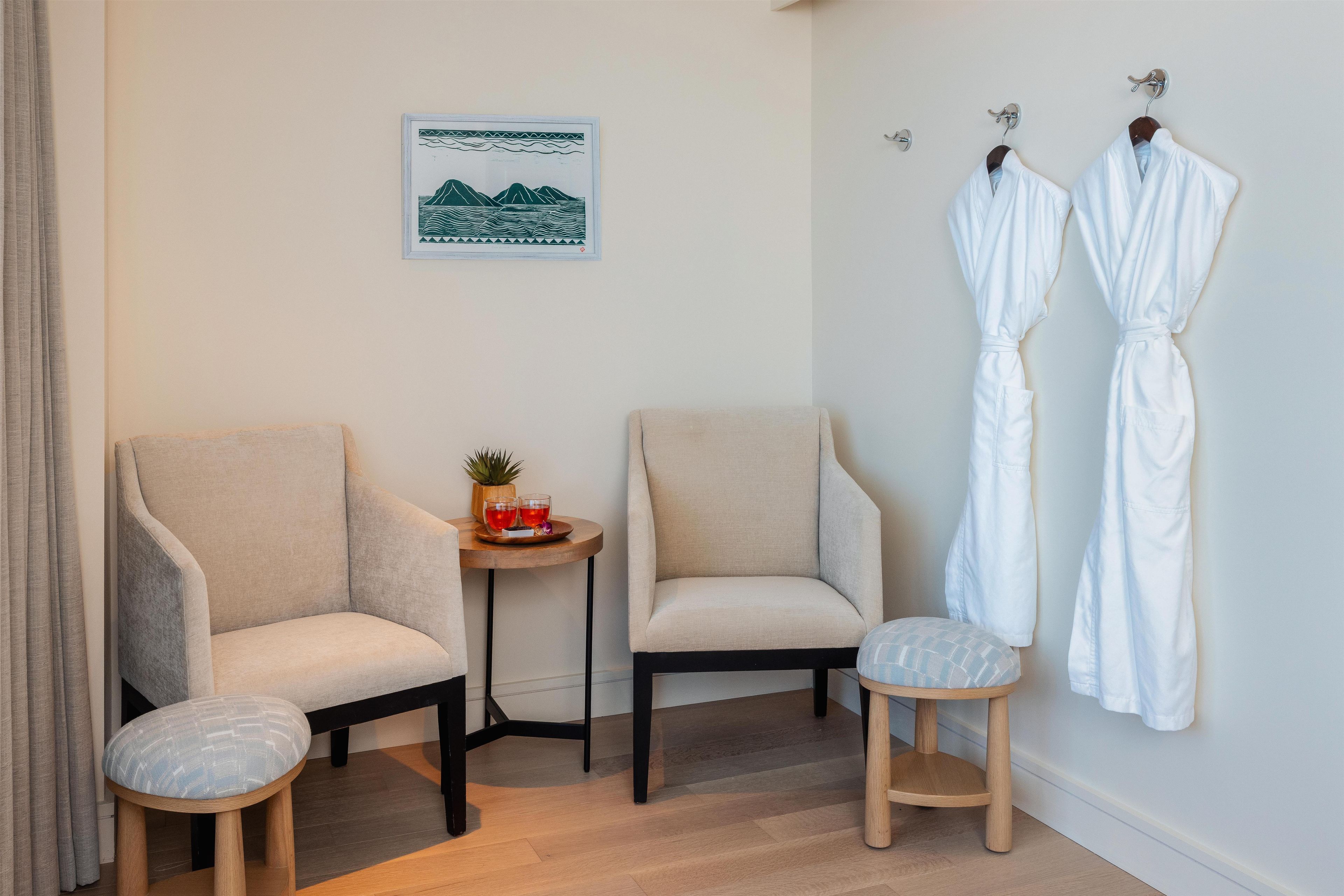 A room with two armchairs, a small round table with a plant and drinks, and two white robes hanging on hooks.