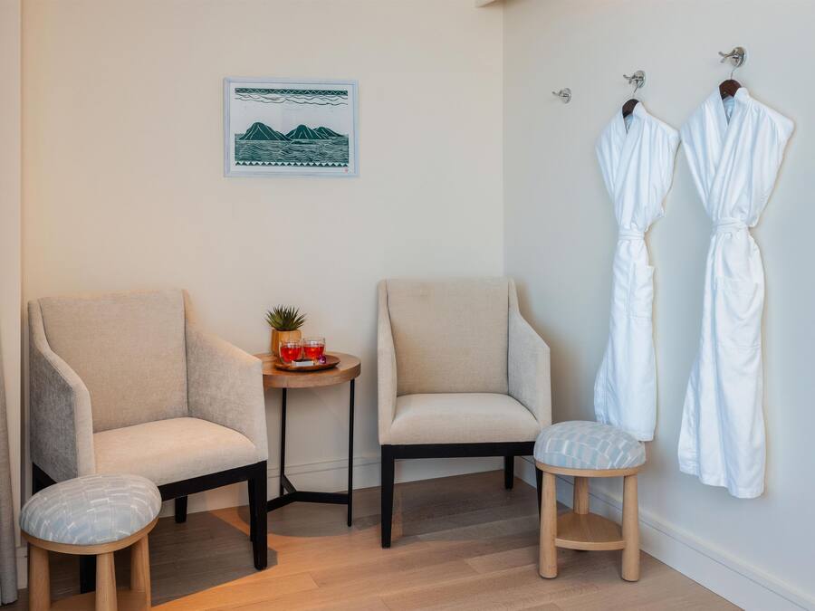 A room with two armchairs, a small round table with a plant and drinks, and two white robes hanging on hooks.