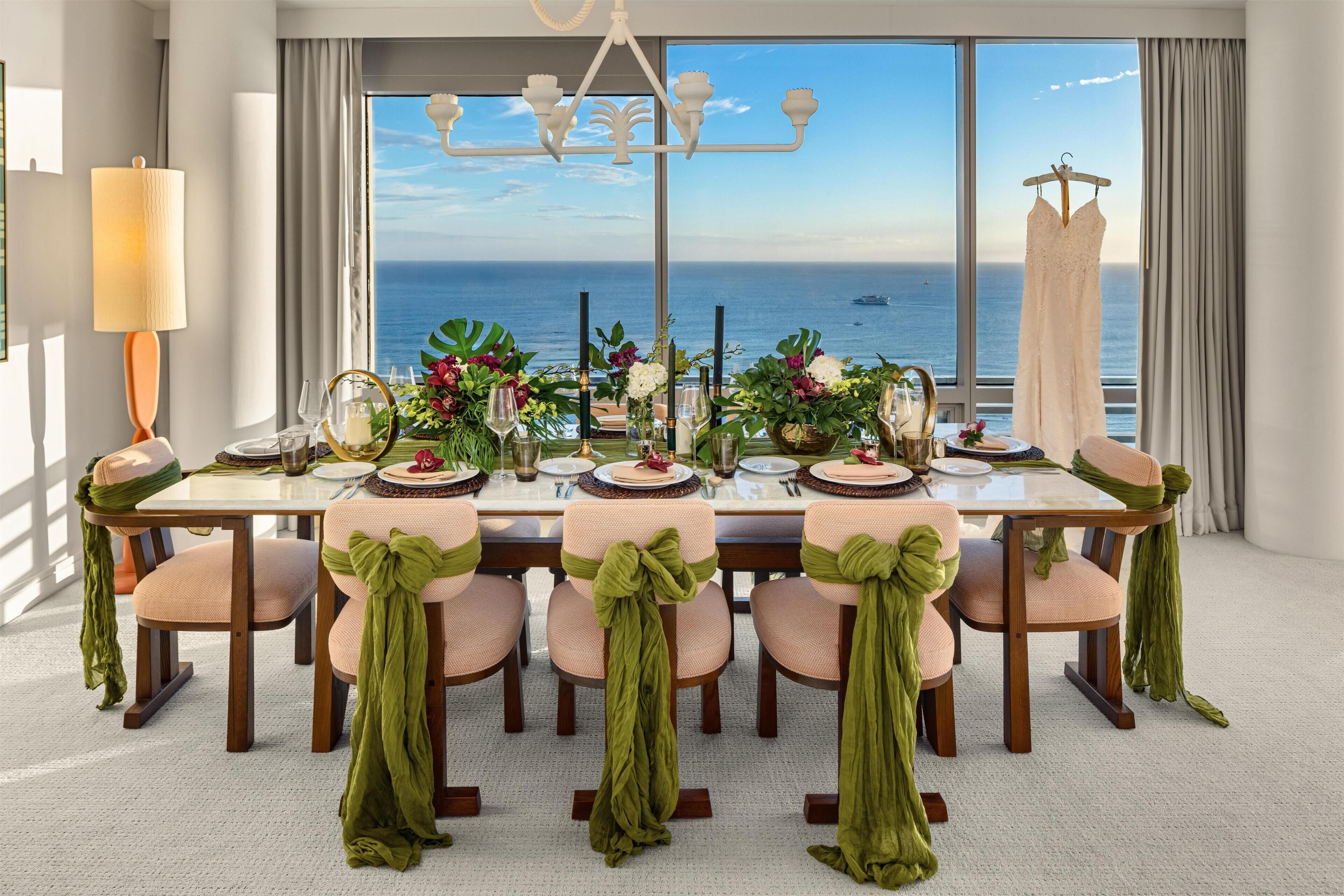 A dining table set for six with chairs, a chandelier, and a view of the ocean.