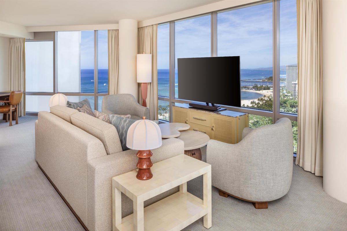 Een moderne hotelkamer met een groot raam, een flatscreen televisie, een bank, een stoel, een lamp en een kleine tafel.