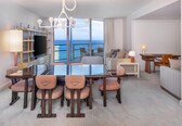 A hotel room with a dining area, a TV, a sofa, a desk, and a view of the ocean.
