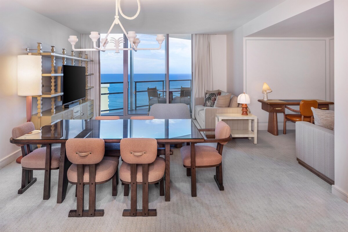 A hotel room with a dining area, a TV, a sofa, a desk, and a view of the ocean.