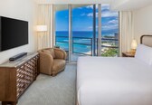 A hotel room with a large bed, a flat-screen TV, a wooden dresser, a comfortable armchair, and a balcony with a view of the ocean and beach.