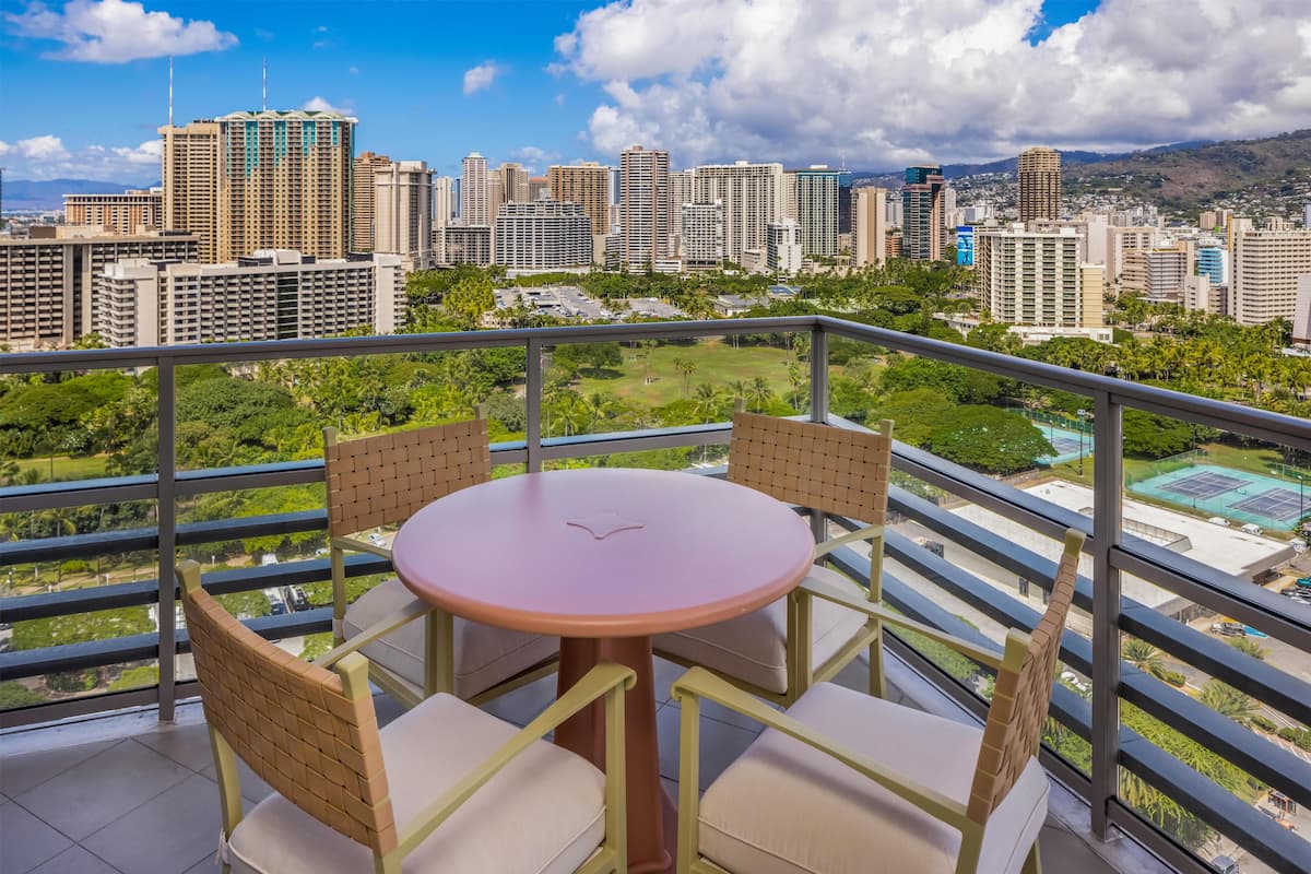 Een balkon met een ronde tafel en vier stoelen, met uitzicht op een stadsgezicht met hoogbouw en een groen park.