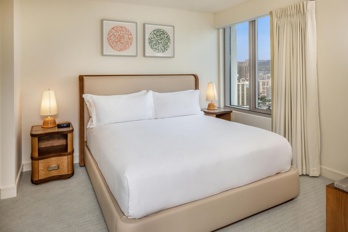 A hotel room with a large bed, two bedside lamps, a nightstand, and a window with a city view.