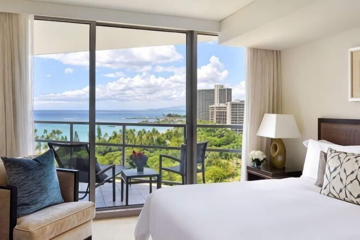A hotel room with a large balcony, a bed, a nightstand, and a view of the ocean and buildings.