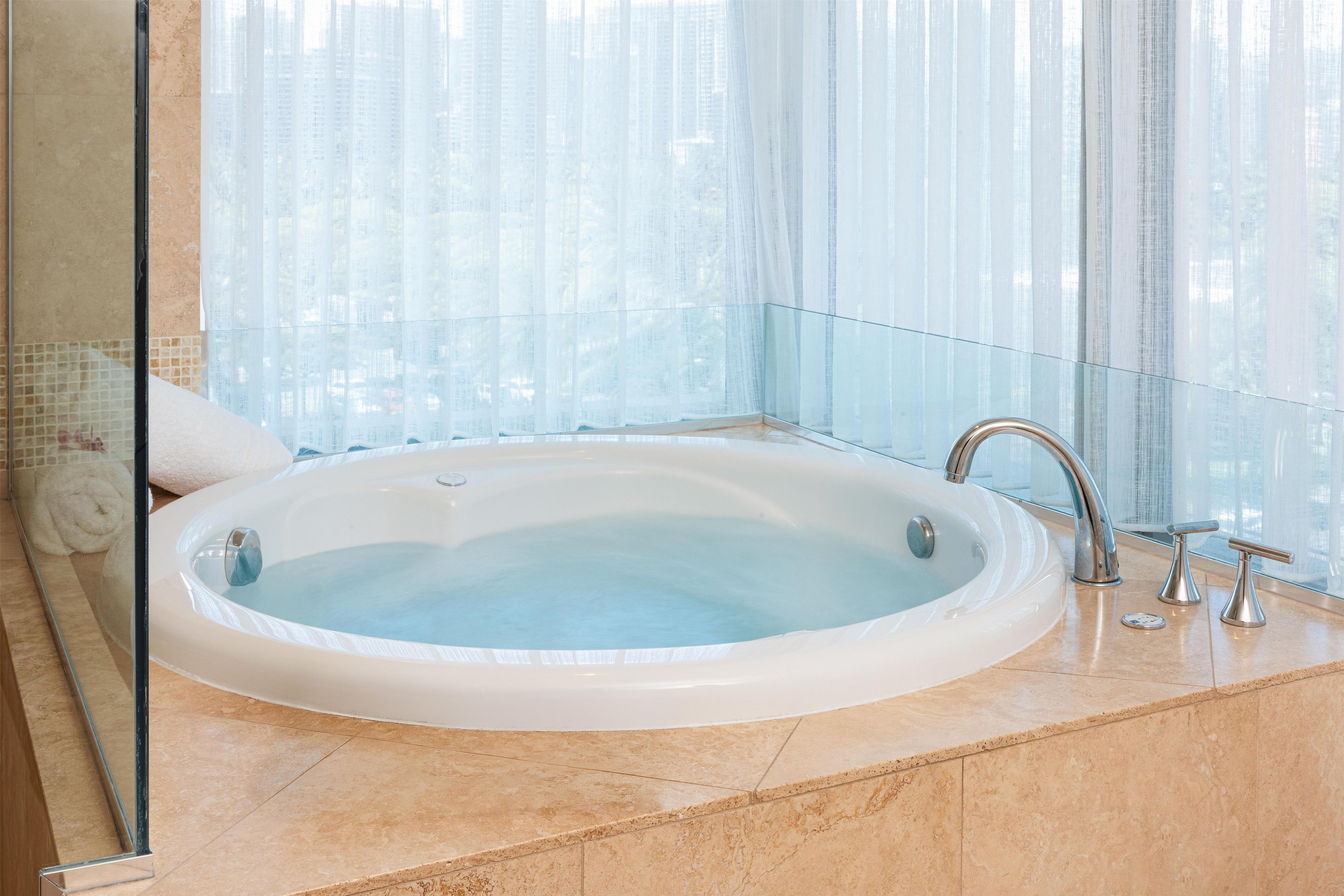 A large, oval-shaped hot tub in a modern bathroom with a glass partition and a view of tall buildings.