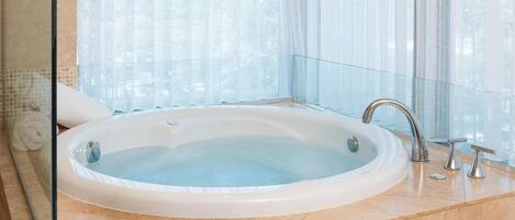 A large, oval-shaped hot tub in a modern bathroom with a glass partition and a view of tall buildings.