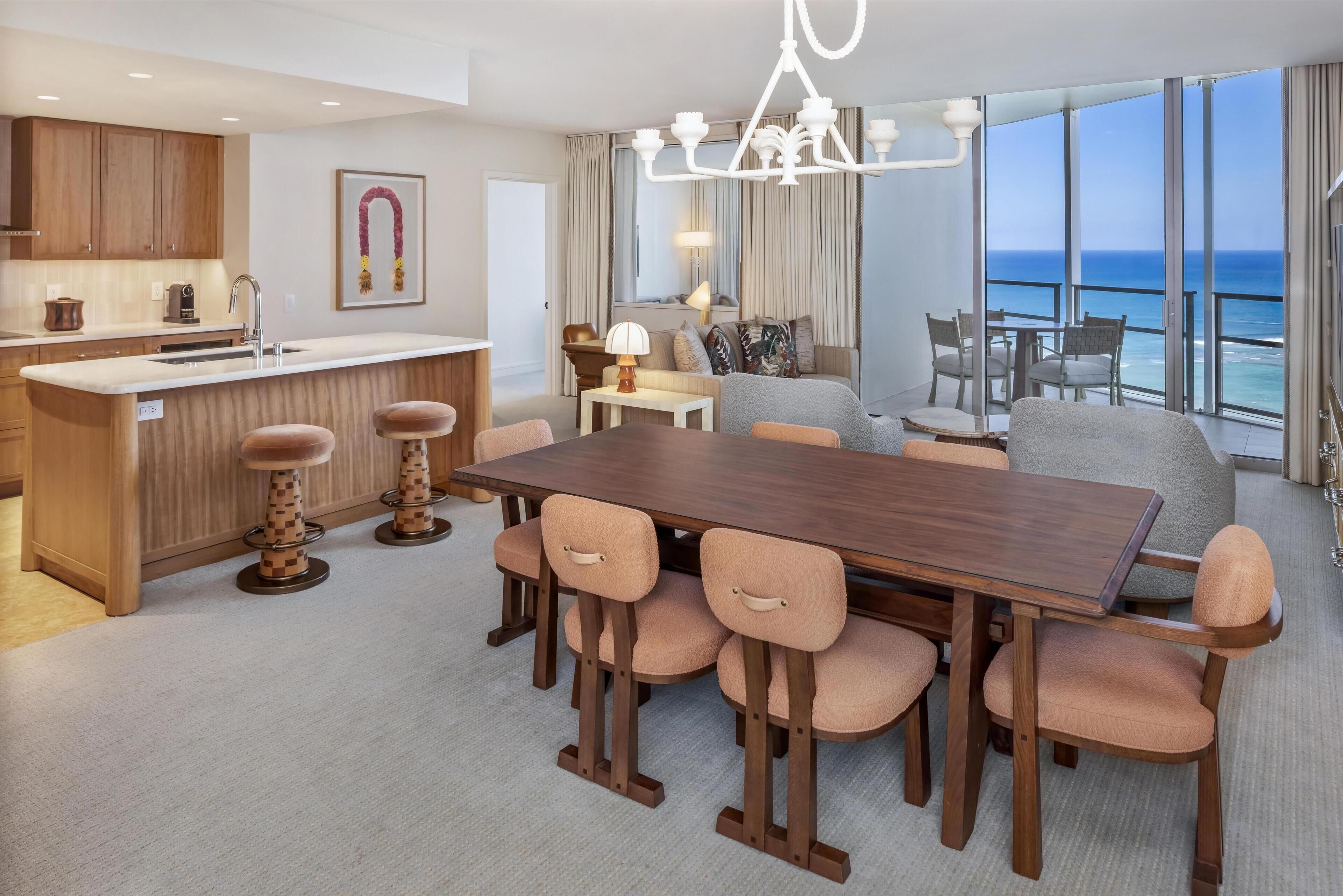 A modern hotel room with a dining area, kitchen, and a balcony with a view of the ocean.