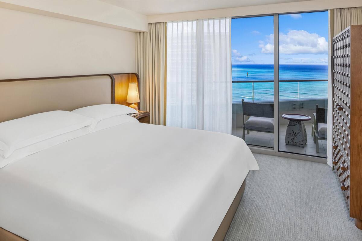 Een hotelkamer met een groot bed, een nachtkastje met lamp, een balkon met uitzicht op de oceaan en een kleine tafel met stoelen.
