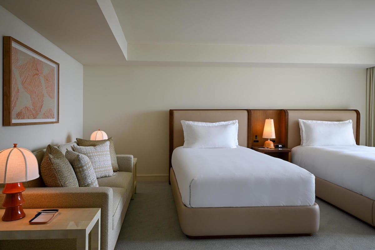 Een hotelkamer met twee bedden, een bank, een lamp, een ingelijst schilderij aan de muur en een nachtkastje met een telefoon.
