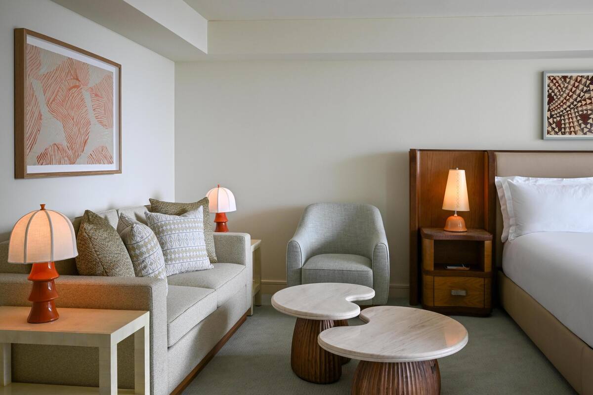 Een hotelkamer met een bank, fauteuil, twee nachtkastjes met lampen, een bed en kunstwerken aan de muur.