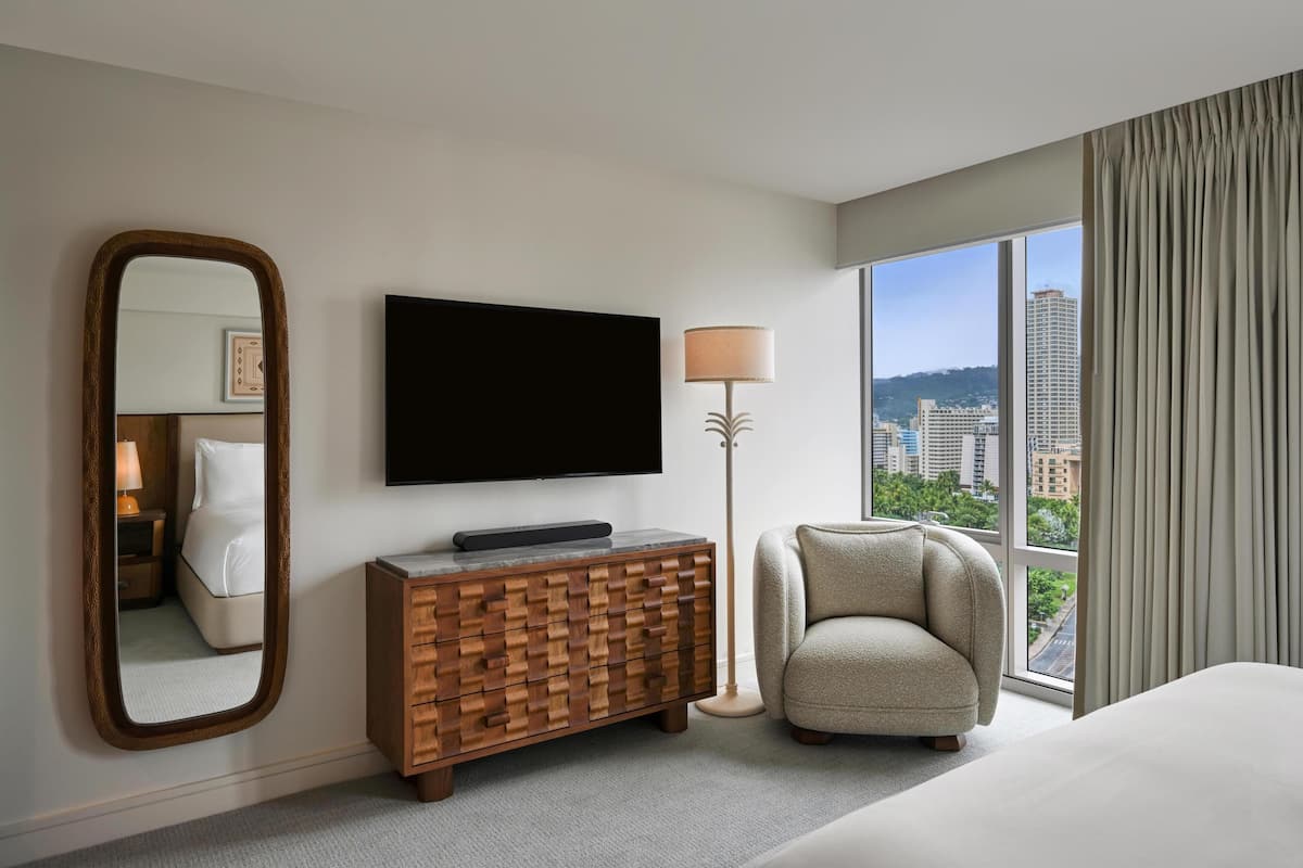 Hotelkamer met een grote spiegel, een flatscreen-tv, een houten dressoir, een lamp, een fauteuil en een groot raam met uitzicht op de stad.