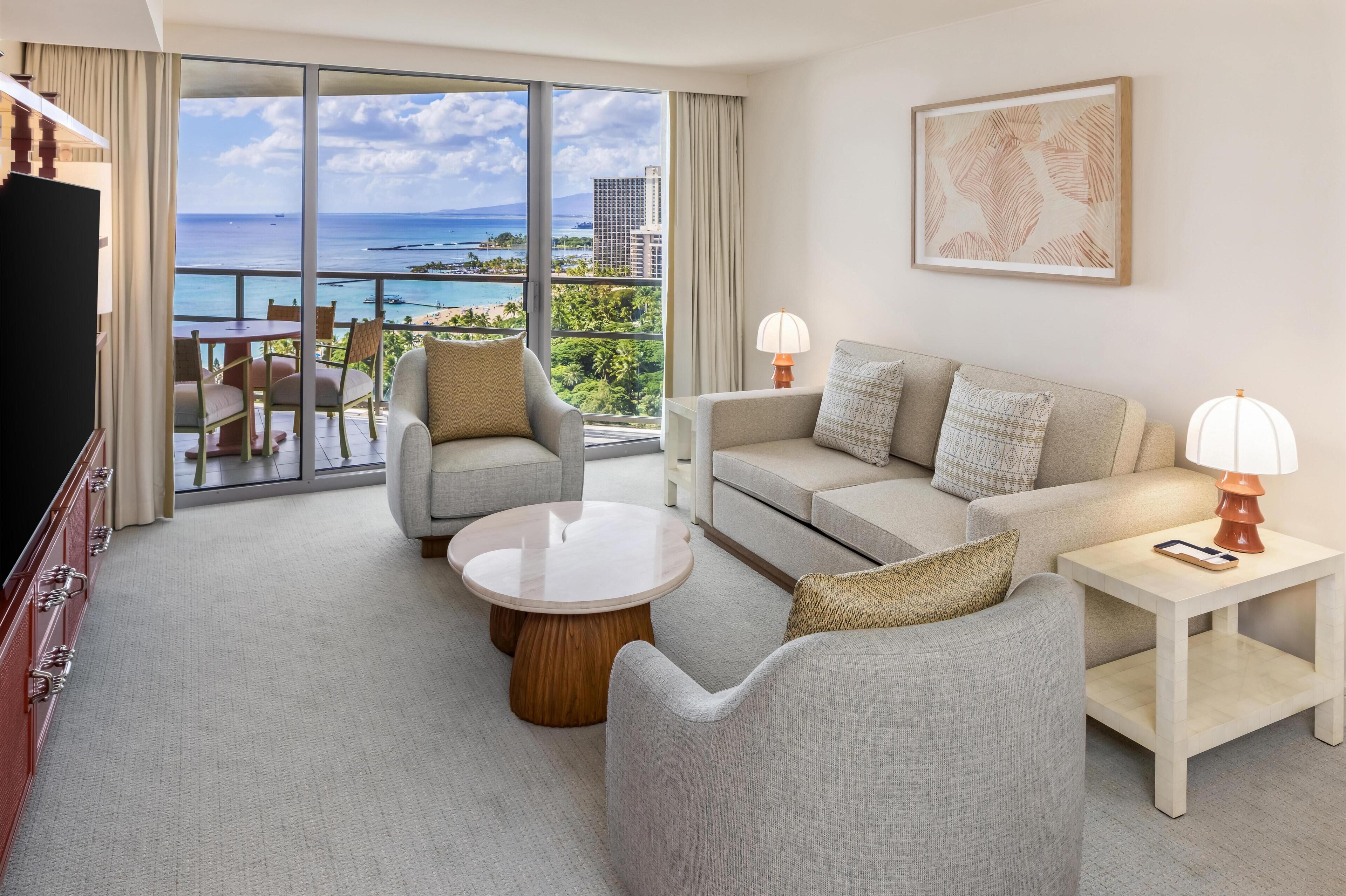 A modern hotel room with a balcony overlooking a scenic ocean view, featuring a sofa, armchairs, a coffee table, and a TV.