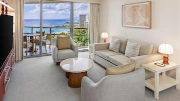 A modern hotel room with a balcony overlooking a scenic ocean view, featuring a sofa, armchairs, a coffee table, and a TV.