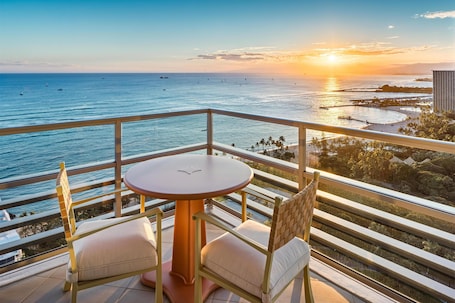 A balcony with a table and chairs, overlooking a coastal view with boats and a sunset.