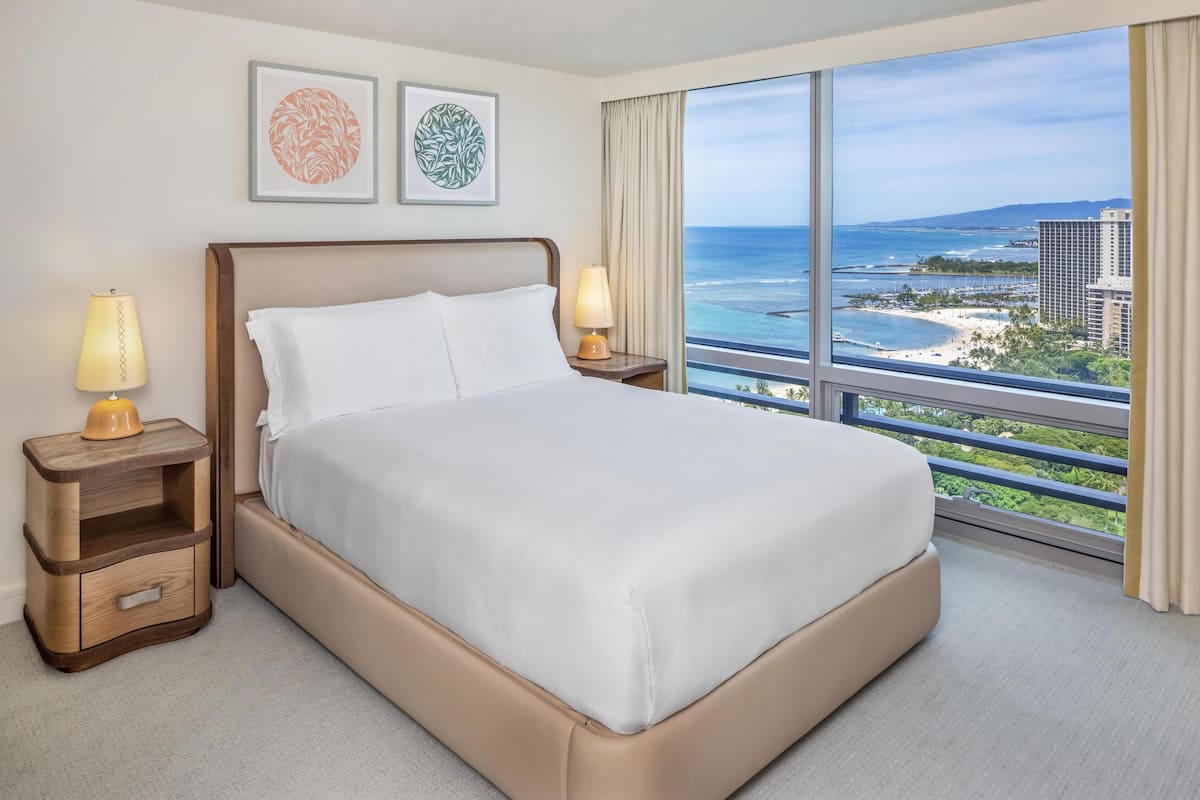 Hotelkamer met een groot bed, twee nachtkastjes en uitzicht op strand en stadsbeeld.