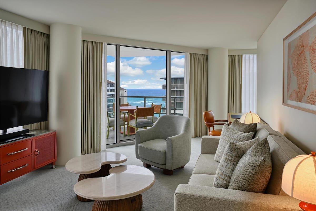Een hotelkamer met een televisie, een bank, een stoel, een tafel en een balkon met uitzicht op de oceaan.