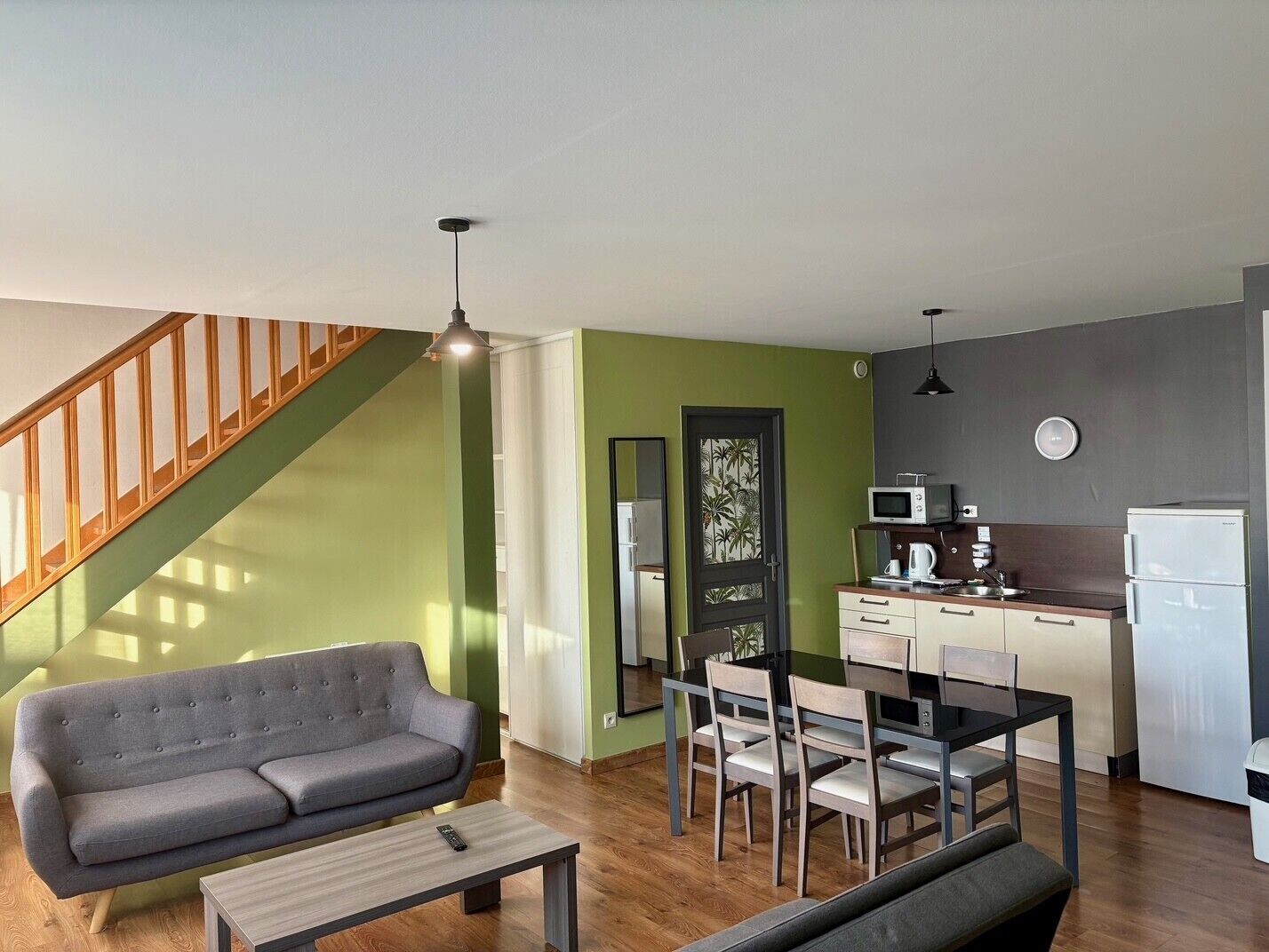Ein modernes Wohnzimmer mit einem grauen Sofa, einem Holztisch und einem Essbereich mit einem schwarzen Tisch und vier Stühlen. Im Hintergrund ist eine Treppe mit Holzgeländern zu sehen.