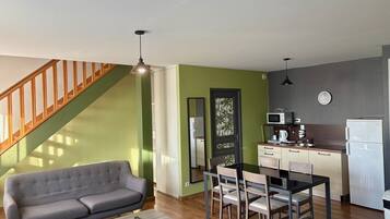 A modern living room with a grey sofa, a wooden coffee table, and a dining area with a black table and four chairs. A staircase with wooden railings is visible in the background.