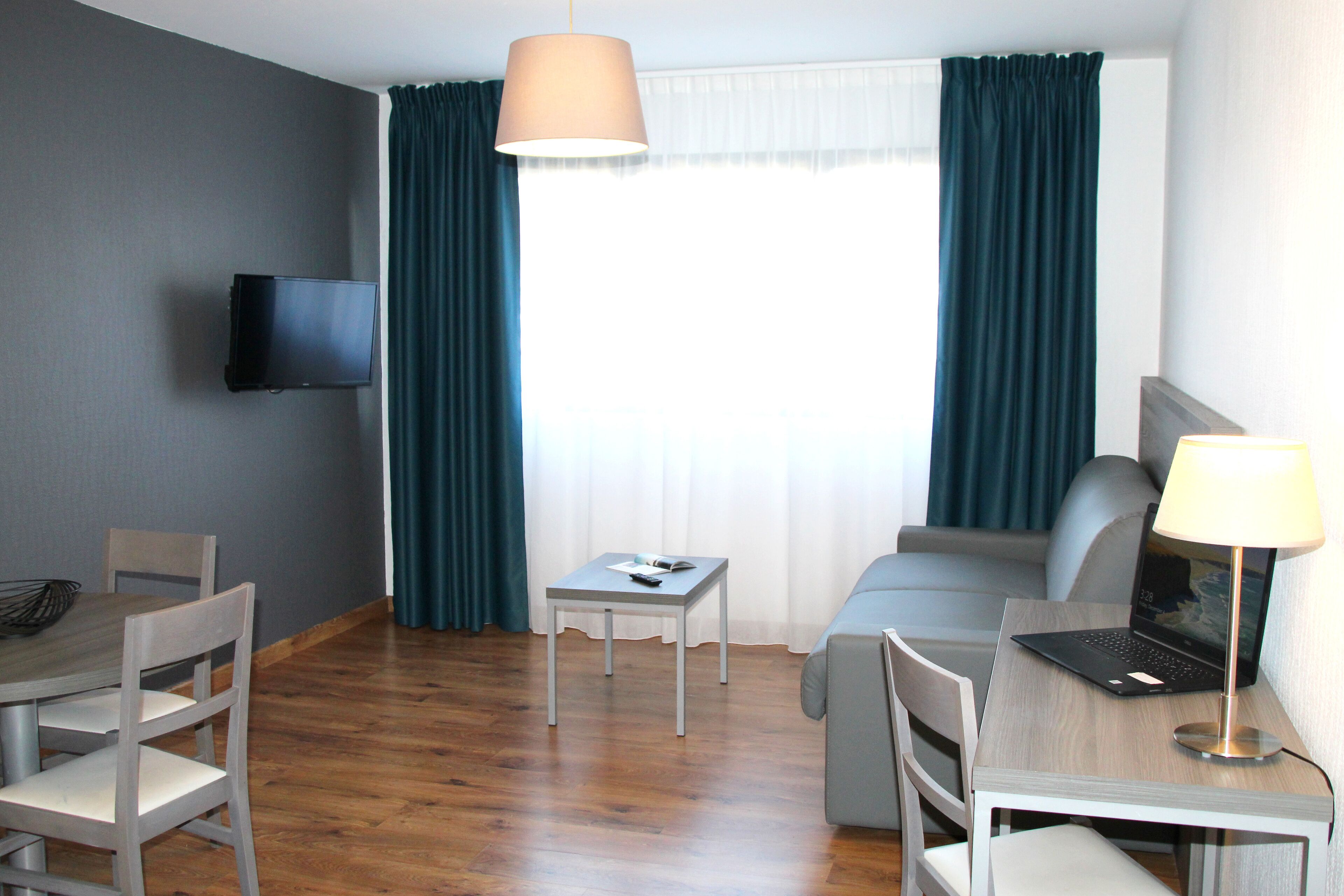 Ein Hotelzimmer mit Holzboden, einem Schreibtisch mit Laptop, einem an der Wand befestigten Fernseher, einer Couch und einem Esstisch mit Stühlen.
