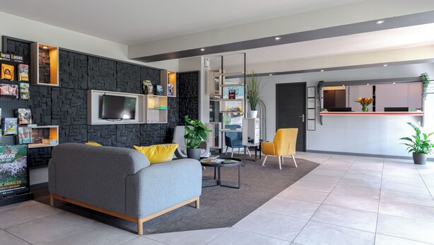 Een moderne hotellobby met een grijze bank, een klein bijzettafeltje en een gele fauteuil. Er is een receptiebalie met een plant en een bloempot op de vloer.
