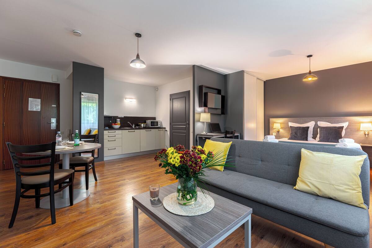 Una sala moderna con un sofá gris, una mesa de comedor con sillas, una mesa de centro con un jarrón de flores y una cocina al fondo.