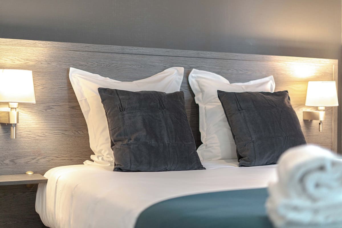 Una cama bien tendida con almohadas blancas y gris oscuro, dos lámparas de noche y un cabecero de madera.