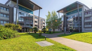 Een modern hotel met een glazen gevel, genaamd 'Zenitude Hotel Residences'. Het heeft een centrale pad, groen en een overdekte ingang.