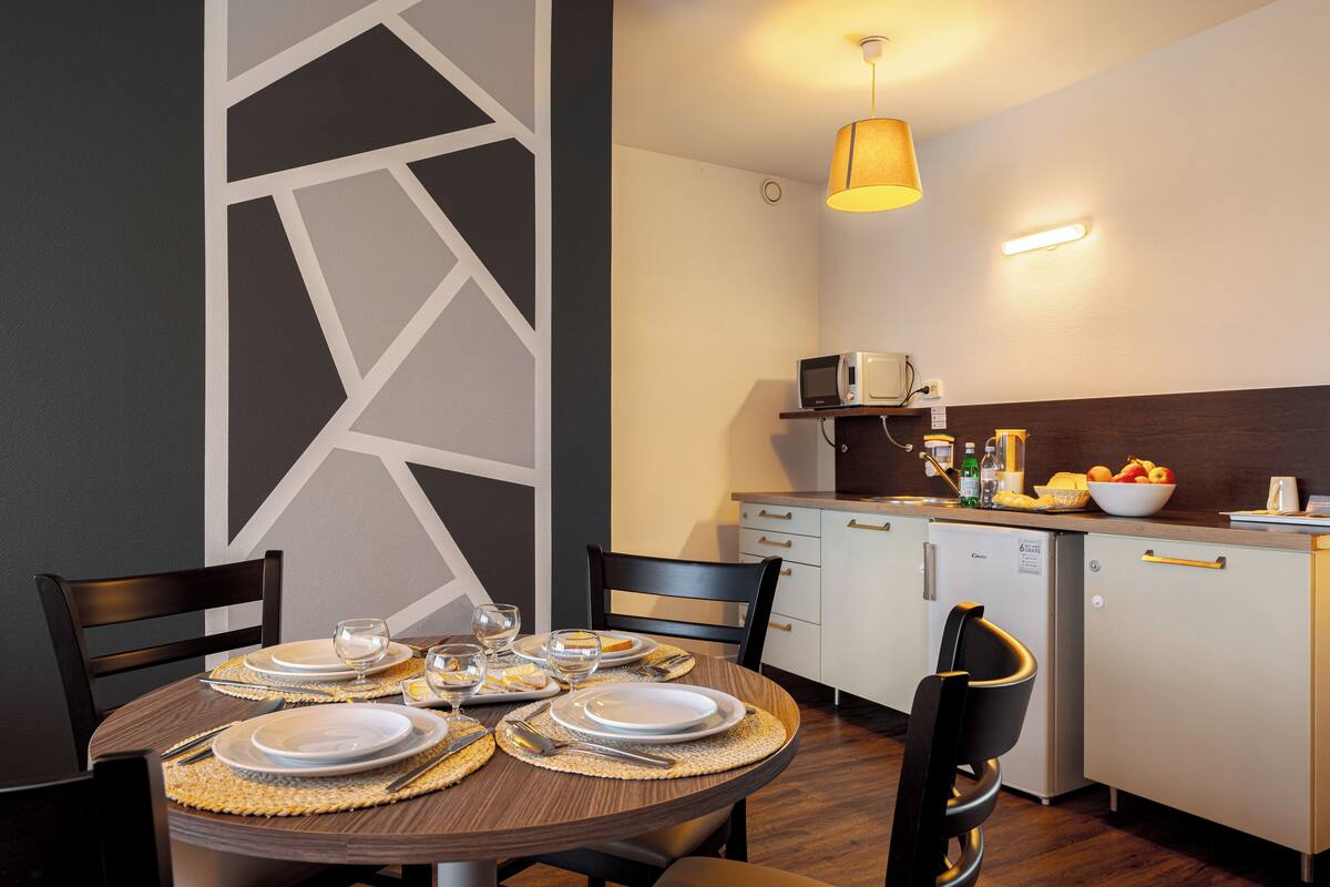 Un comedor con una mesa redonda dispuesta para cuatro personas, con platos, cubiertos y vasos. Se ven un microondas y un refrigerador pequeño al fondo.