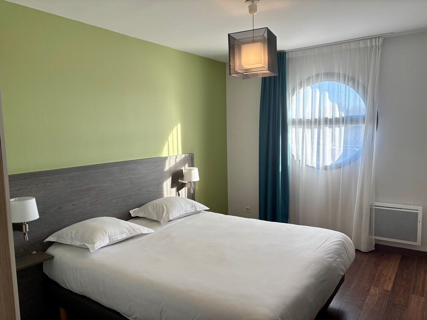 Una camera d'albergo con un letto grande, una testiera, una lampada da comodino, una finestra con tende e una lampada a soffitto.