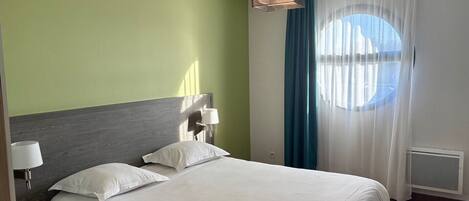 A hotel room with a large bed, a headboard, a bedside lamp, a window with curtains, and a ceiling light fixture.