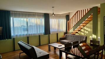 Una sala de estar moderna con sofá, sillón y mesa de comedor. Se observa una escalera con barandilla de madera al fondo.