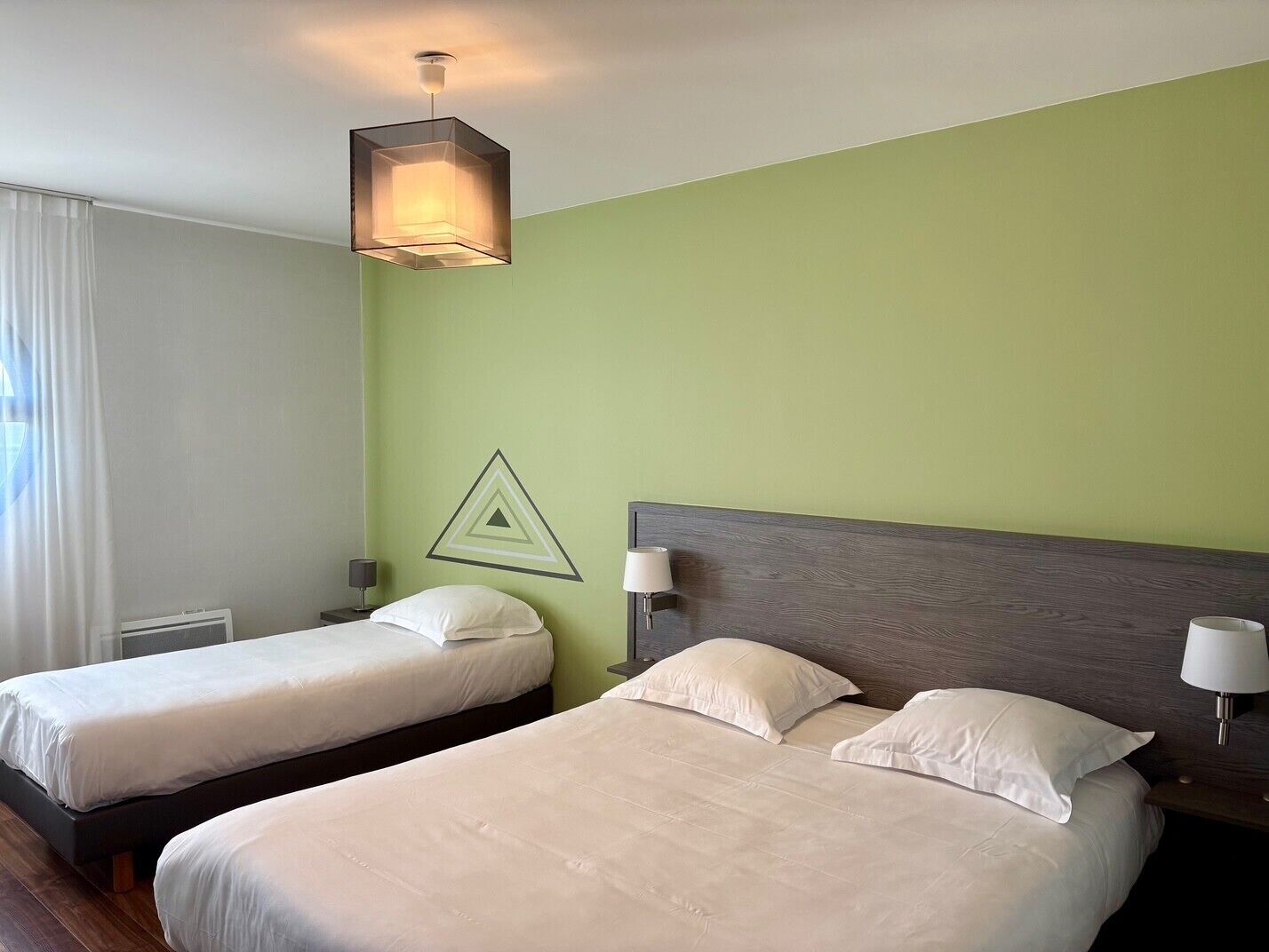 Habitación de hotel con dos camas individuales, una pared de acento verde, un adorno geométrico en la pared y una lámpara colgante.