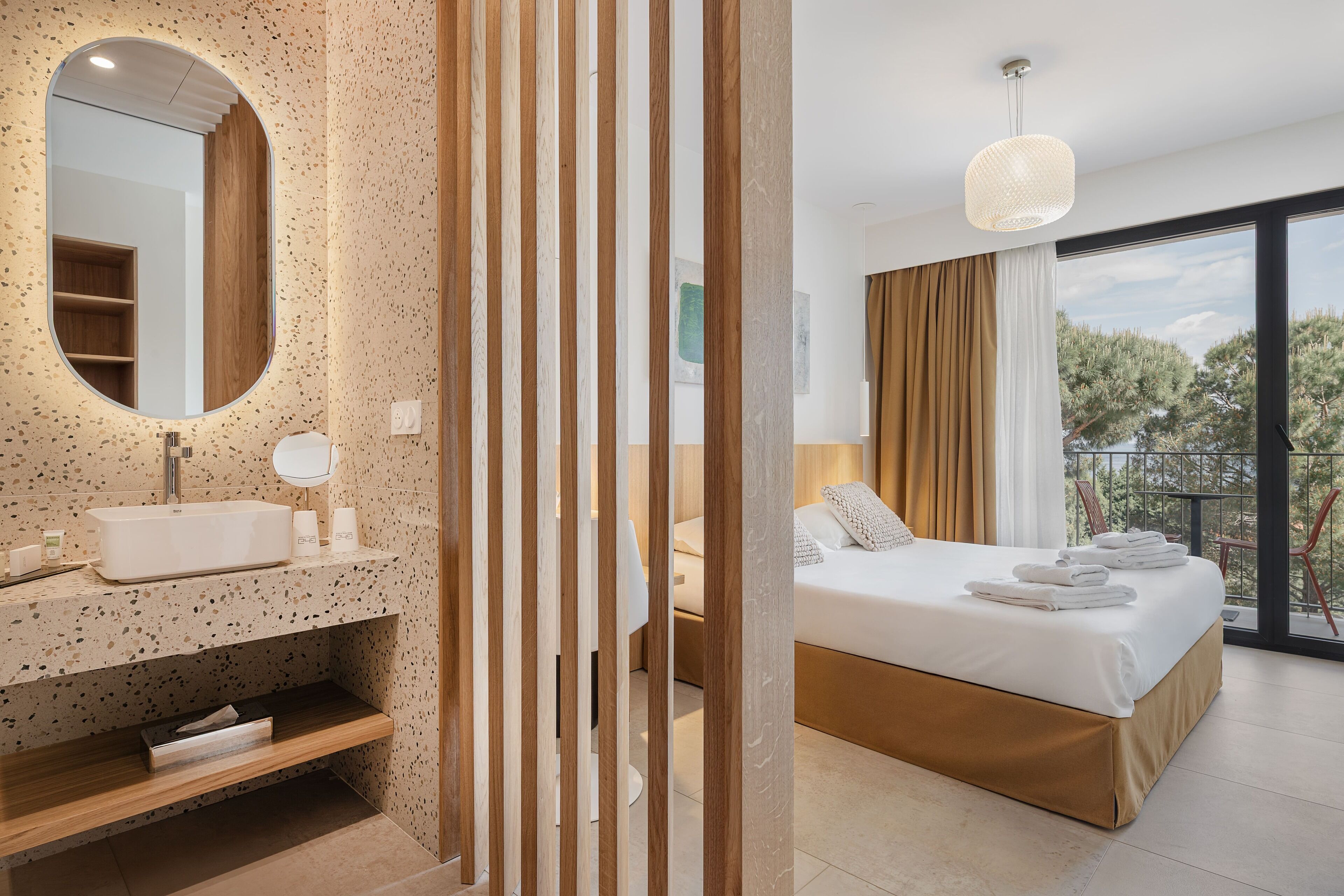 A modern hotel room with a large bed, a bathroom with a freestanding sink, and a balcony with a view of trees.