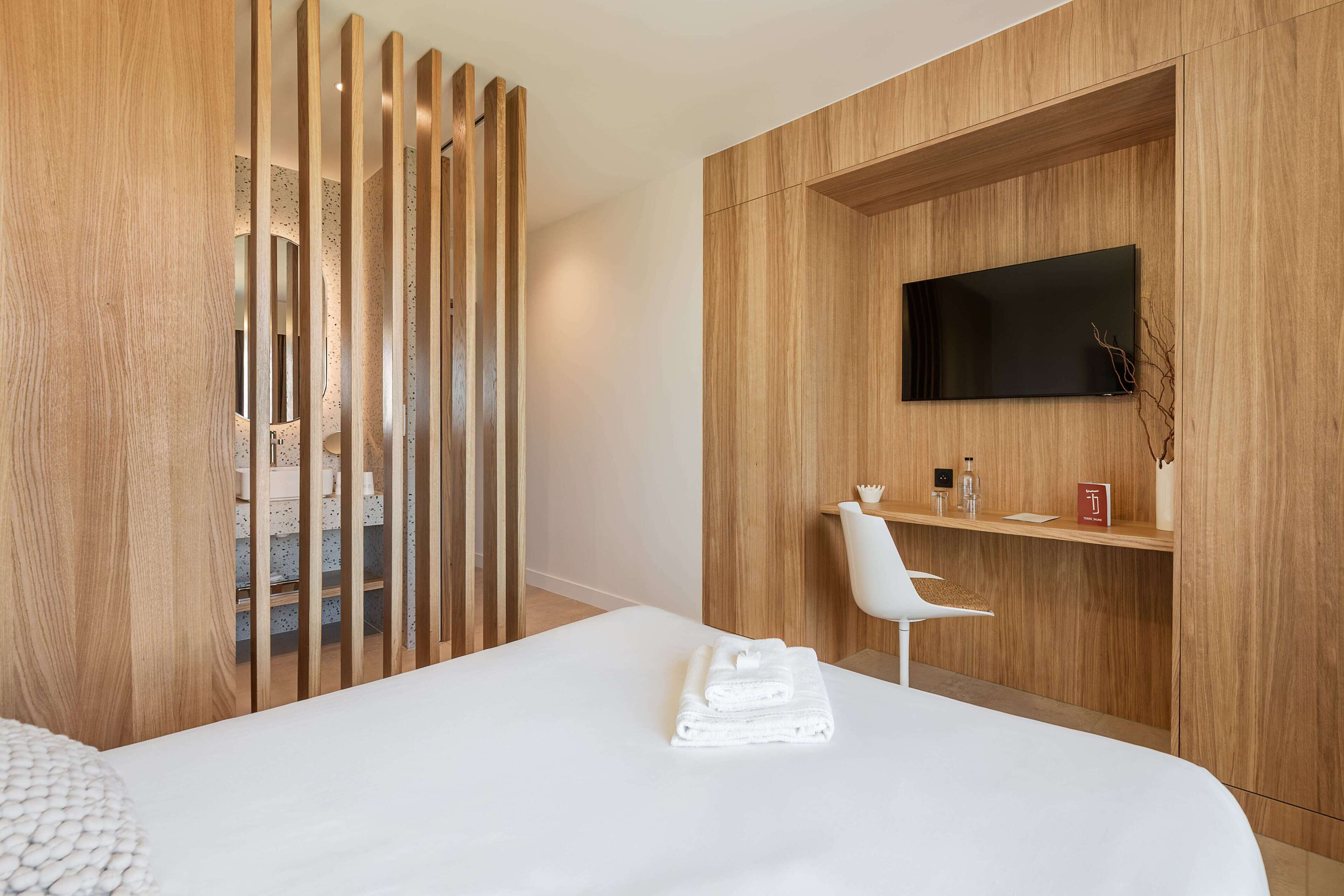 Habitación de hotel moderna con una cama grande, un separador de madera, un televisor montado y una silla blanca.