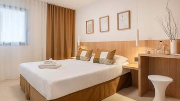 A hotel room with a large bed, a wooden headboard, a bedside table with a lamp, and a small white stool.