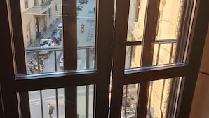 A view through a window with vertical blinds, revealing a street with parked cars and a building with multiple floors and balconies. - Hotel Ester (Florence)