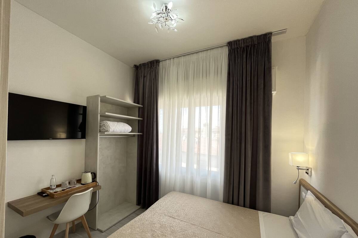 A hotel room with a bed, a desk with a chair, a wardrobe, a TV mounted on the wall, and a window with curtains.
