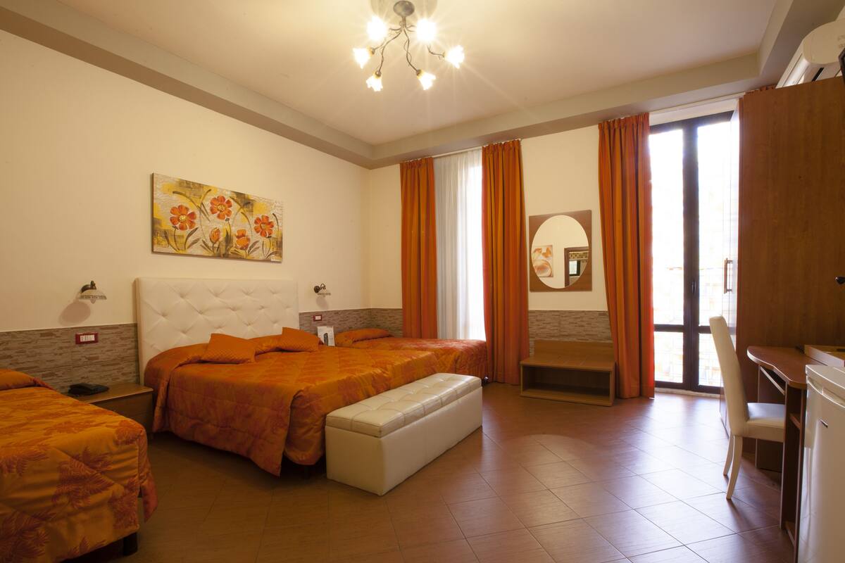 A hotel room with a large bed, a bench, a desk, and a bathroom with a mirror.