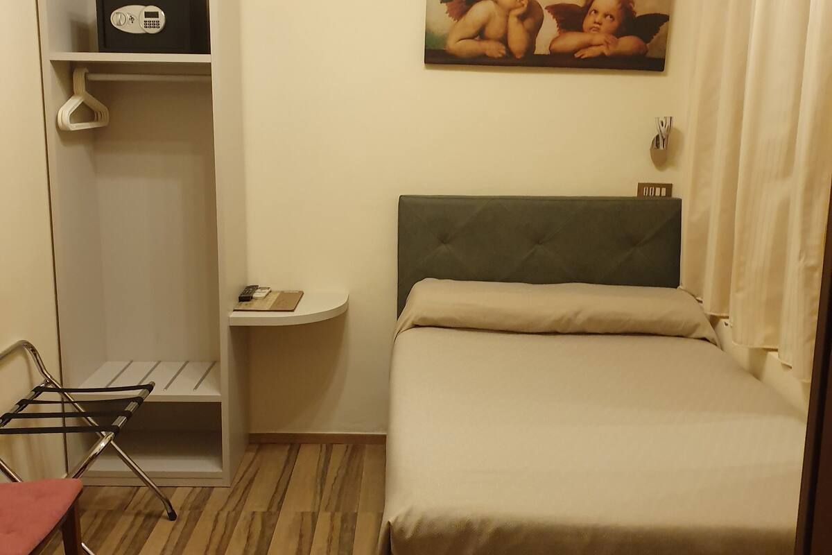 A hotel room with a bed, a small bedside table, a wall-mounted air conditioner, a painting, and a wooden floor.