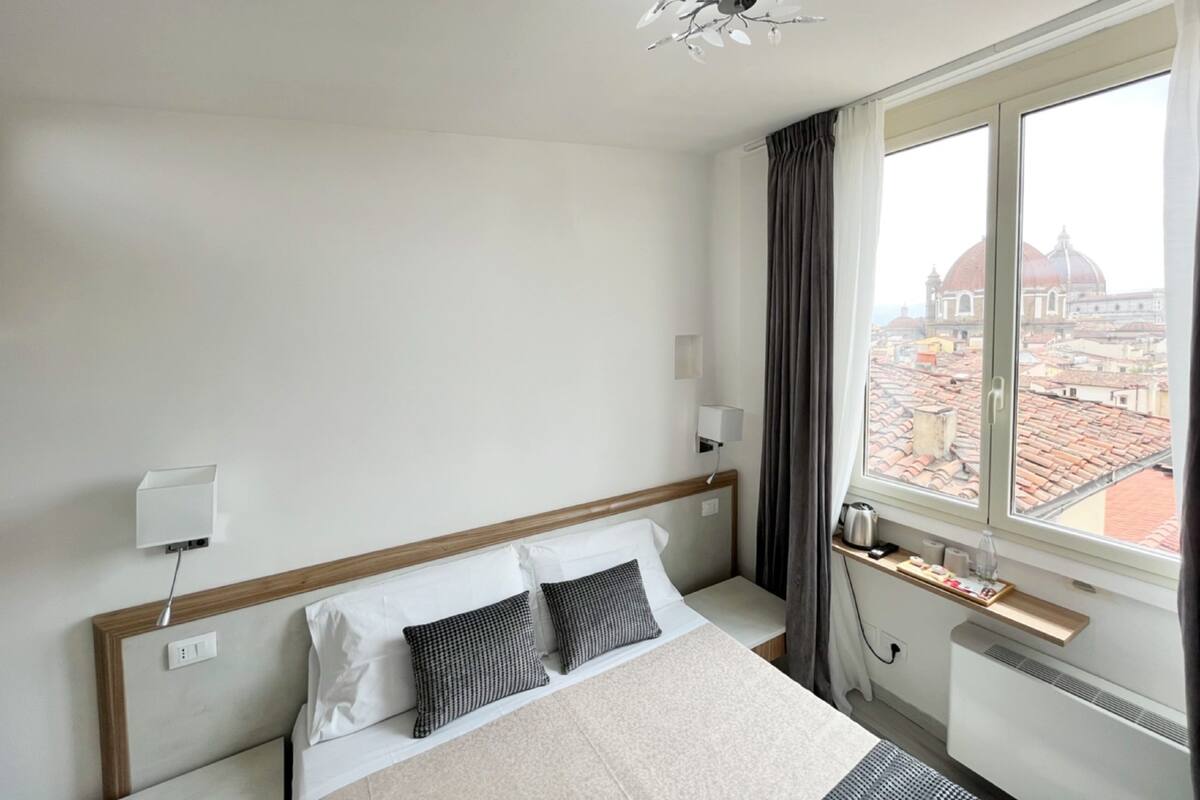 A hotel room with a bed, a window with a city view, a ceiling light, and a power outlet.