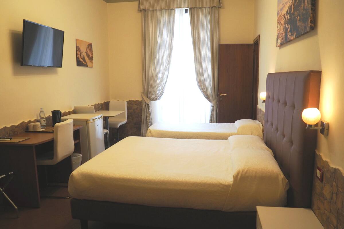 A hotel room with two beds, a desk, a chair, a TV, and a window with curtains.