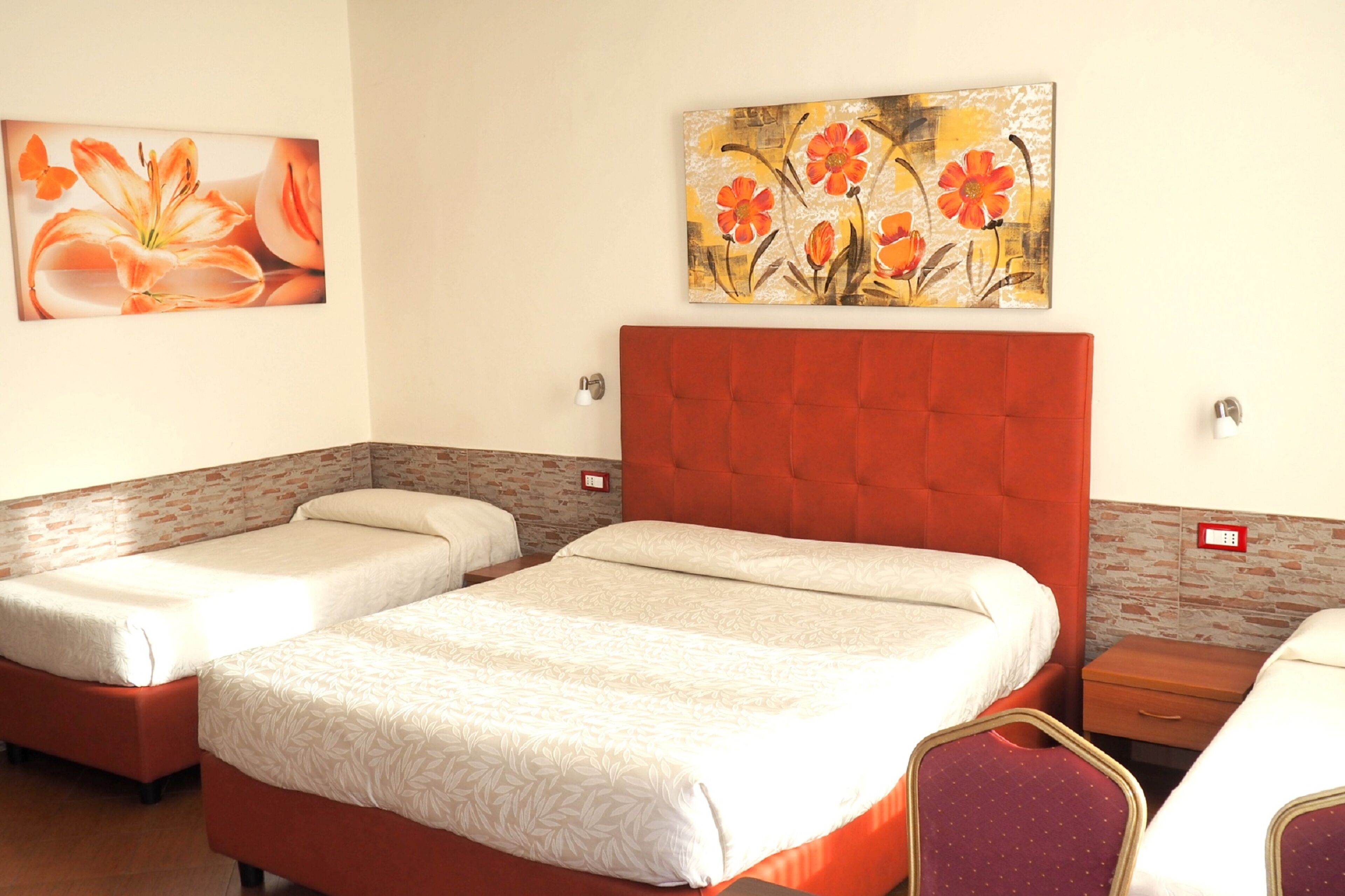 Une chambre d’hôtel avec deux lits simples, une tête de lit rouge, un mur décoré d’un motif en briques et un tableau floral au-dessus des lits.