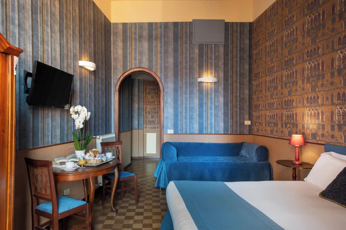 Ein Hotelzimmer mit Schachbrettmuster auf dem Boden, einem Bett mit blauer Bettdecke, einem blauen Sofa, einem kleinen Tisch mit Lampe, einem an der Wand befestigten Fernseher und einem dekorativen Bogen.