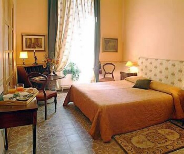 A hotel room with a bed, a desk with a chair, a lamp, and a window with curtains.