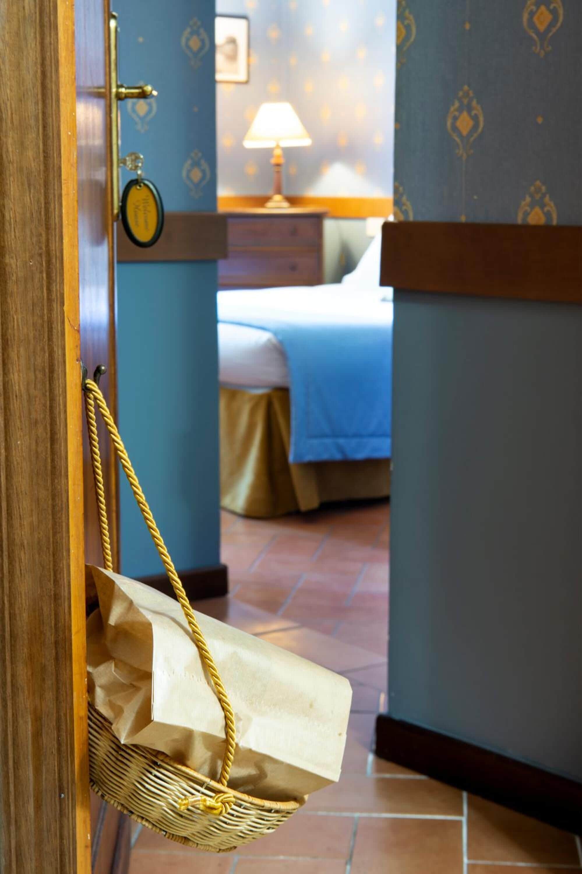 A hotel room with a bed, a nightstand, and a lamp. A wicker basket with a paper bag hangs on the door.